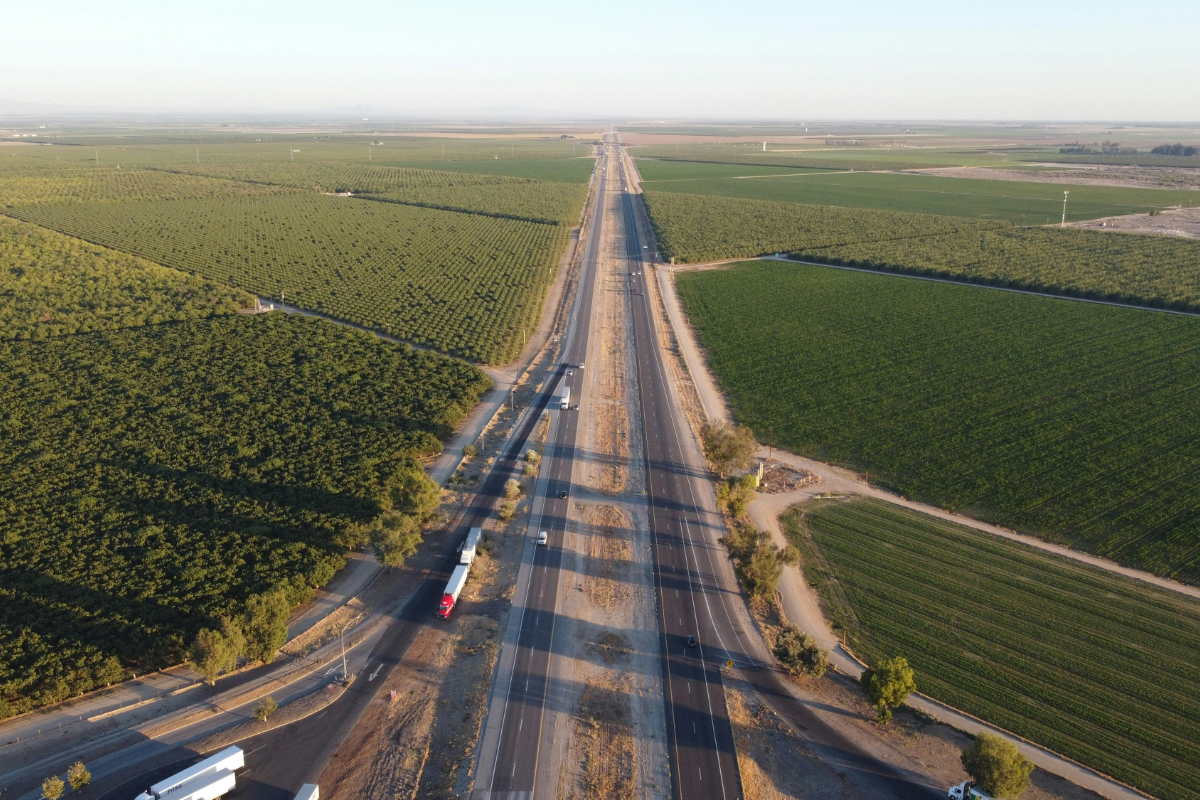 Aerial view of a divided highway running between orchards and green farm fields, with trucks traveling in both directions