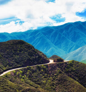 Winding mountain road surrounded by lush green hills with bright blue mountains and clouds in the distance.